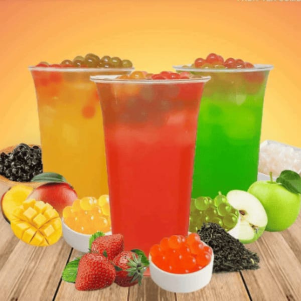 Three colorful fruit teas with popping boba, surrounded by fresh fruit and tea leaves on a wooden surface.